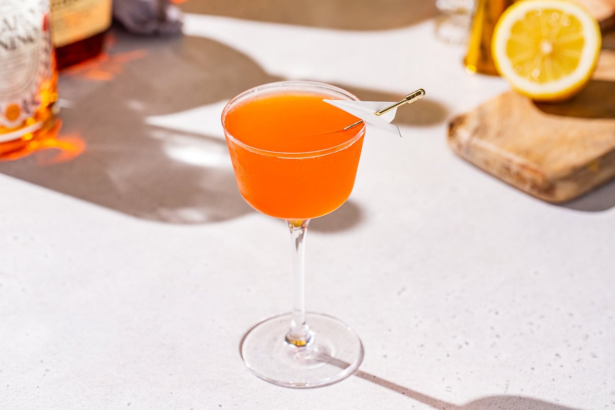 Slightly overhead view of an orange colored Paper Plane cocktail in a coupe glass. The drink is garnished with a mini paper plane on a cocktail pick.