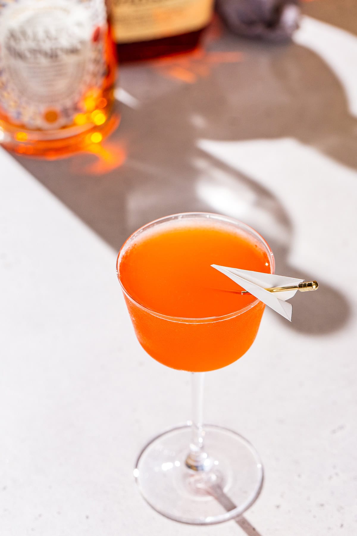 Paper Plane cocktail on a countertop with bottles of liqueur in the background.