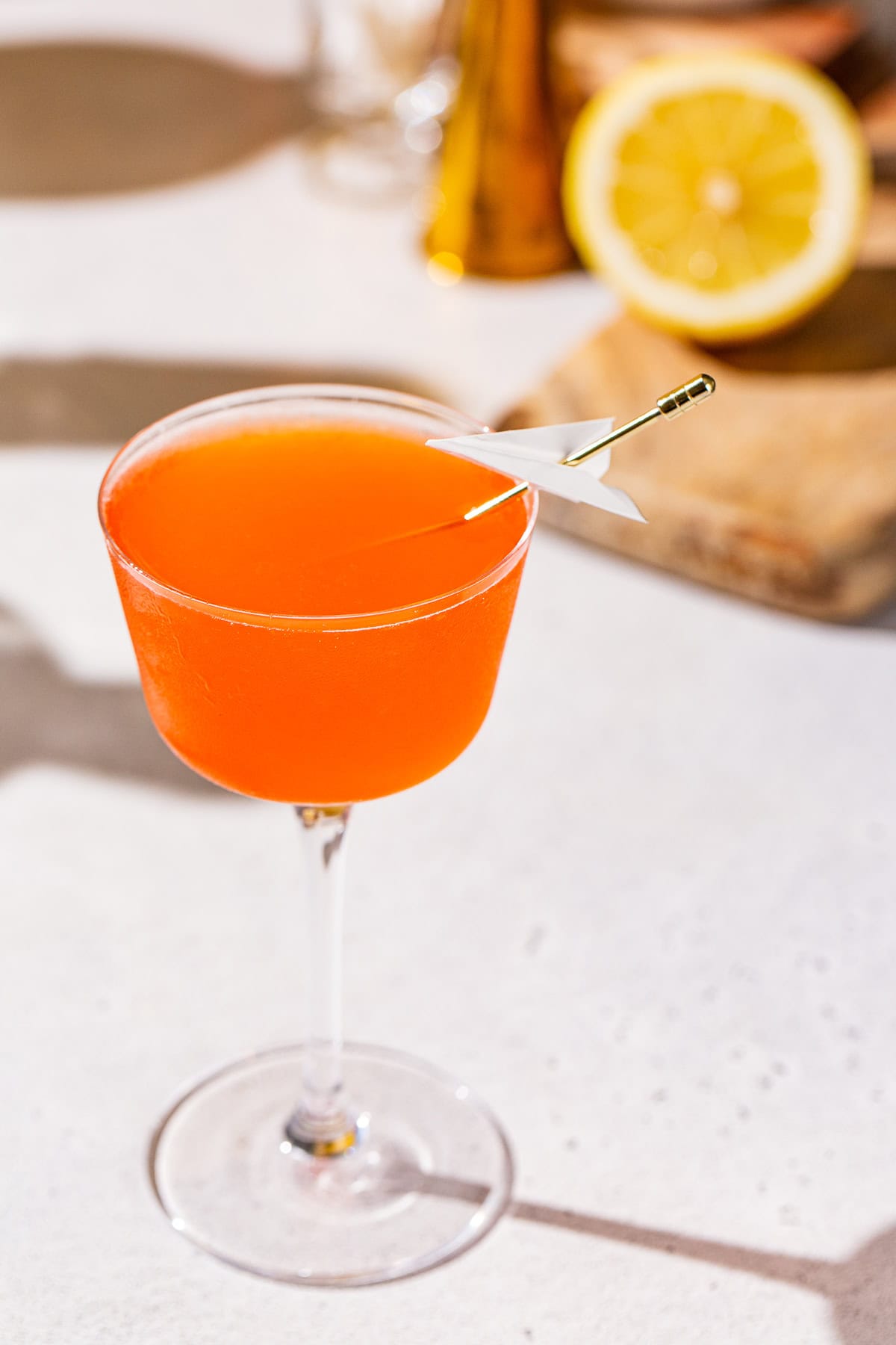 Paper Plane cocktail on a countertop with a cutting board and a lemon in the background.