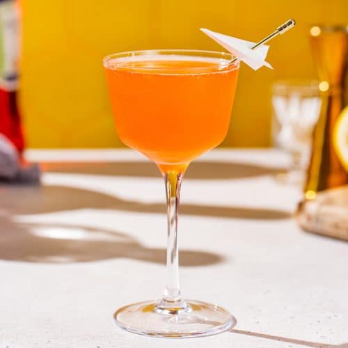 Side view of a Paper Plane cocktail with a jigger and other ingredients in the background.