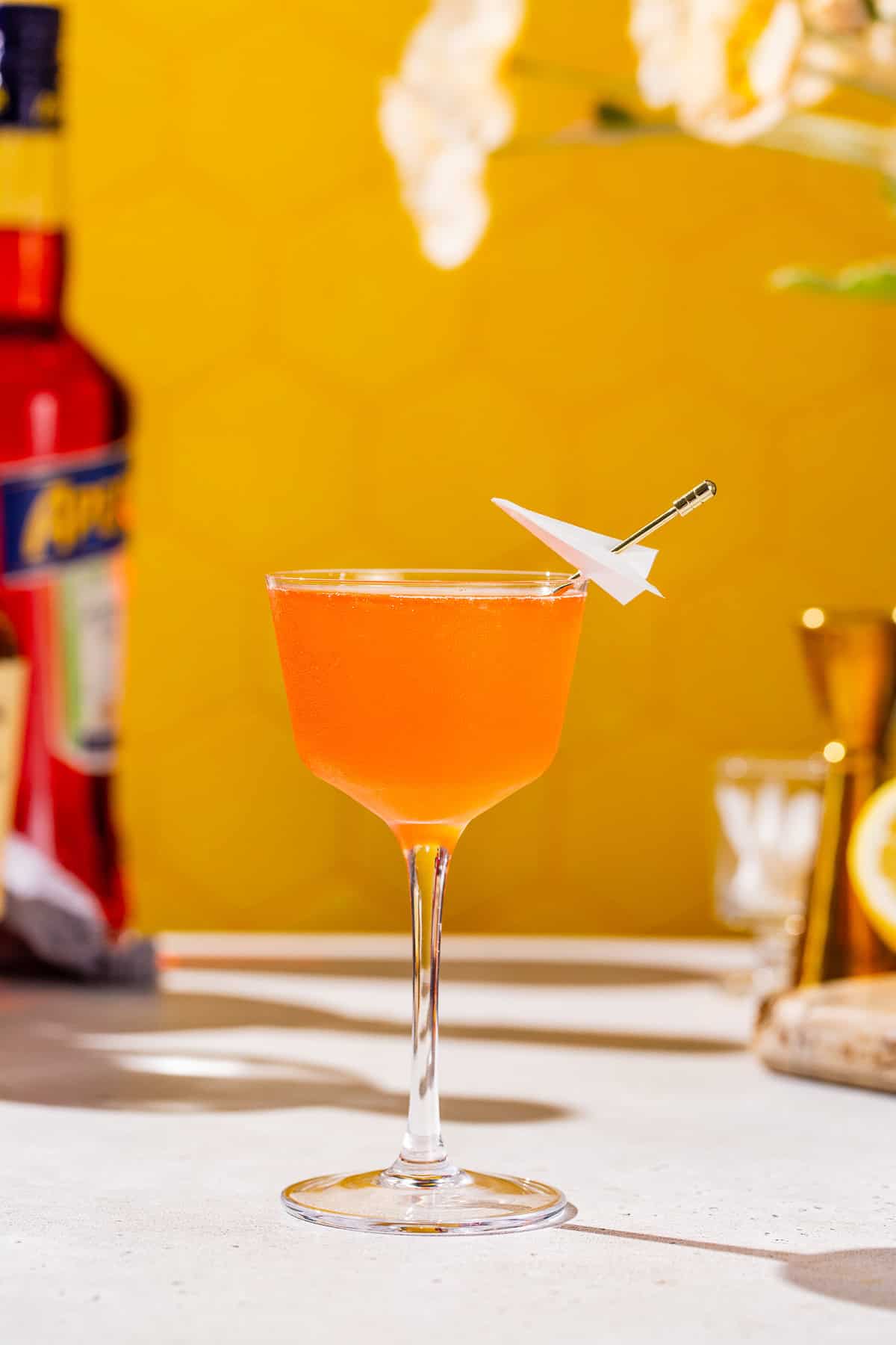 Side view of a Paper Plane cocktail. The drink is bright orange and has a mini paper plane on a cocktail pick for a garnish.