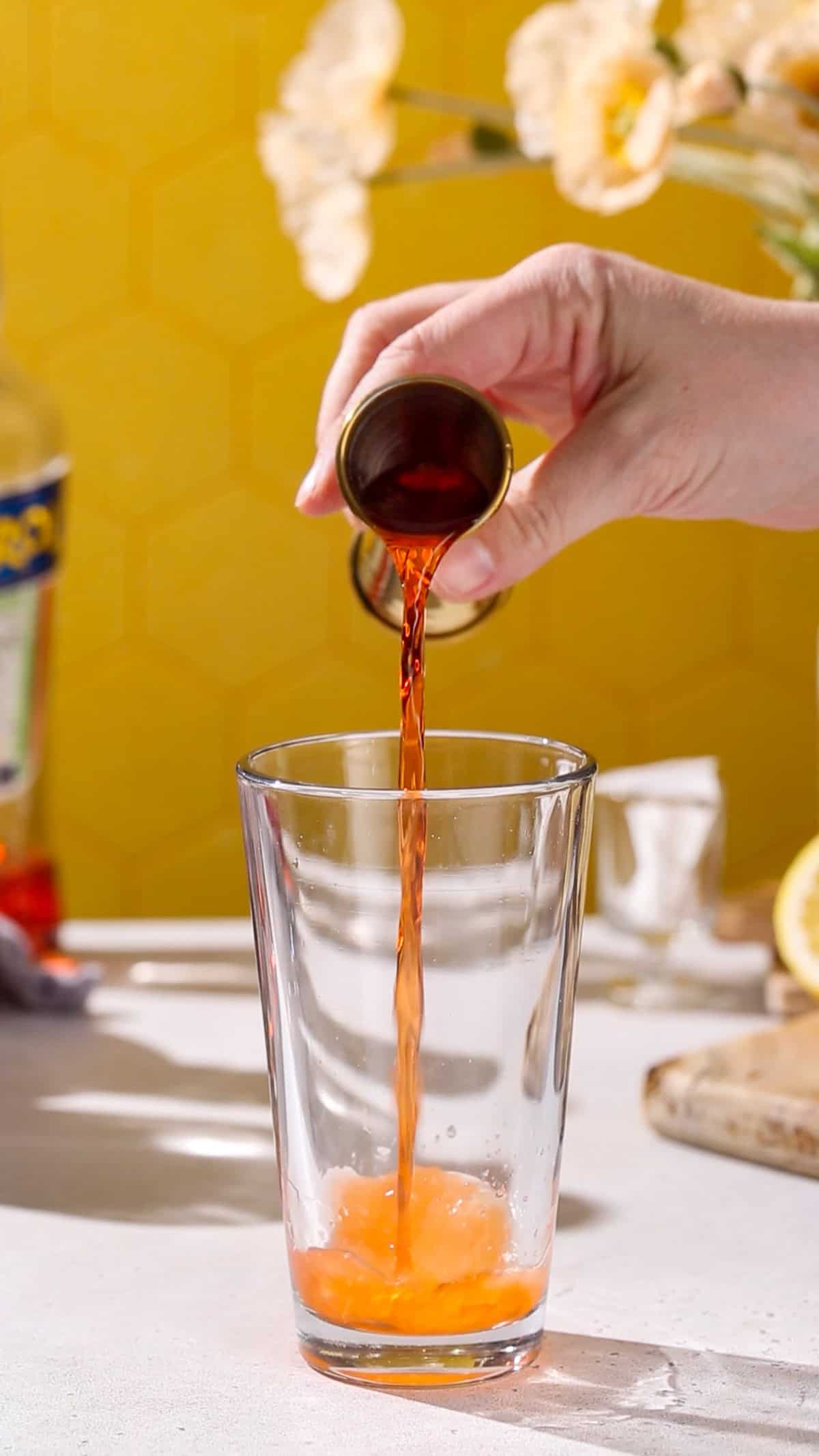 Hand adding aperol liqueur to a cocktail shaker.