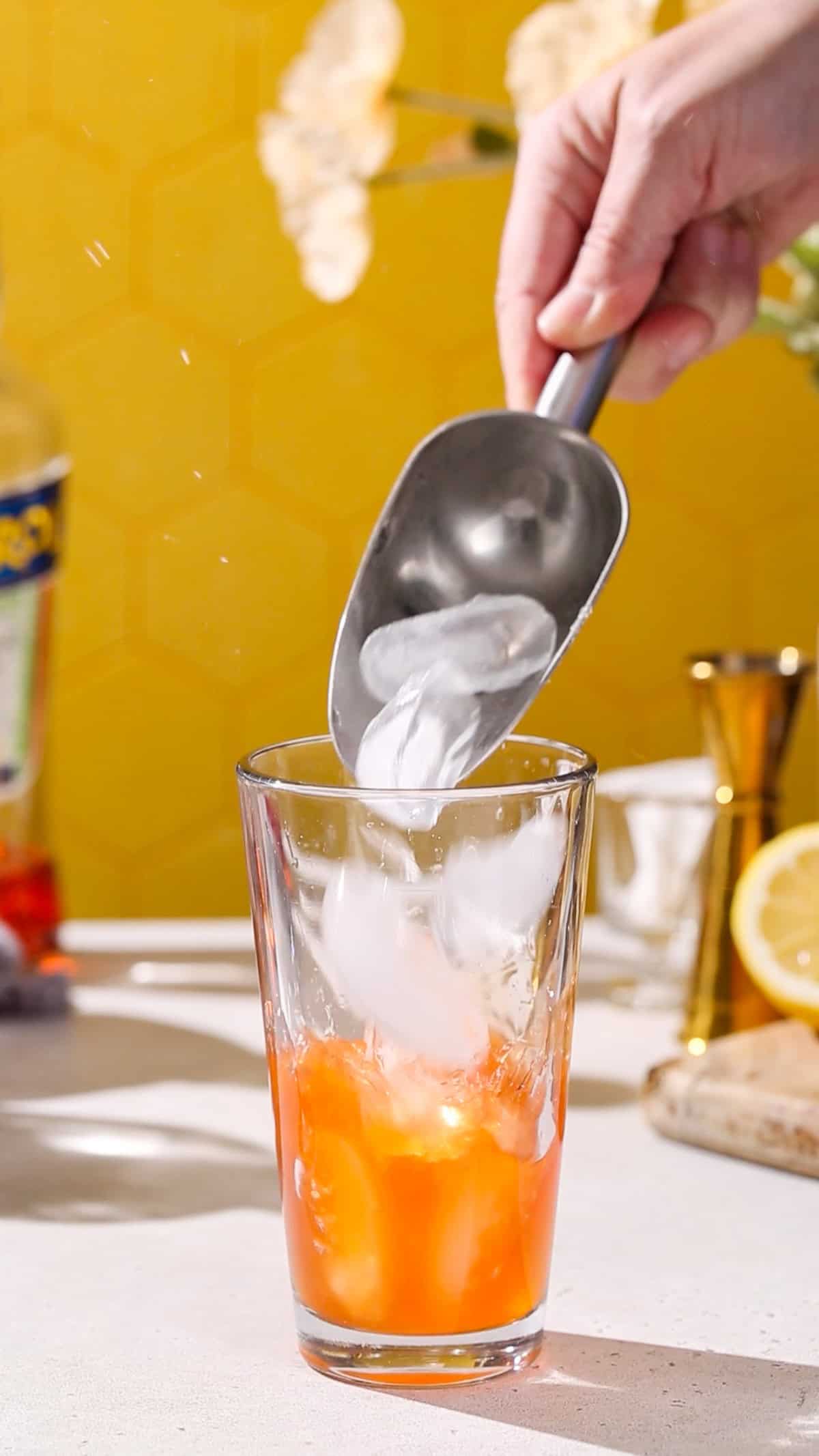 Hand adding ice to a cocktail shaker filled with orange liquid.