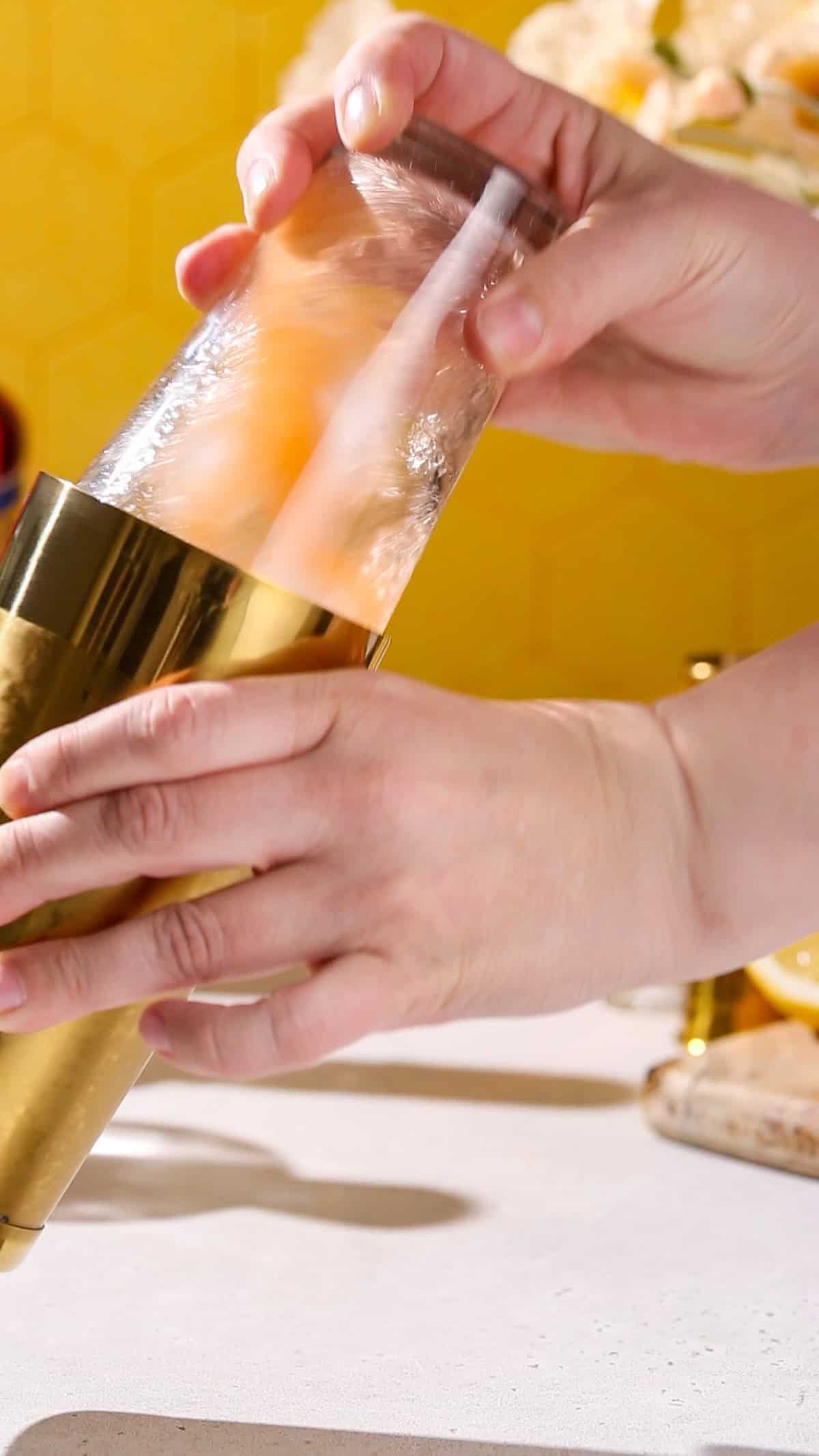 Hands shaking a cocktail shaker filled with ice and orange liquid.