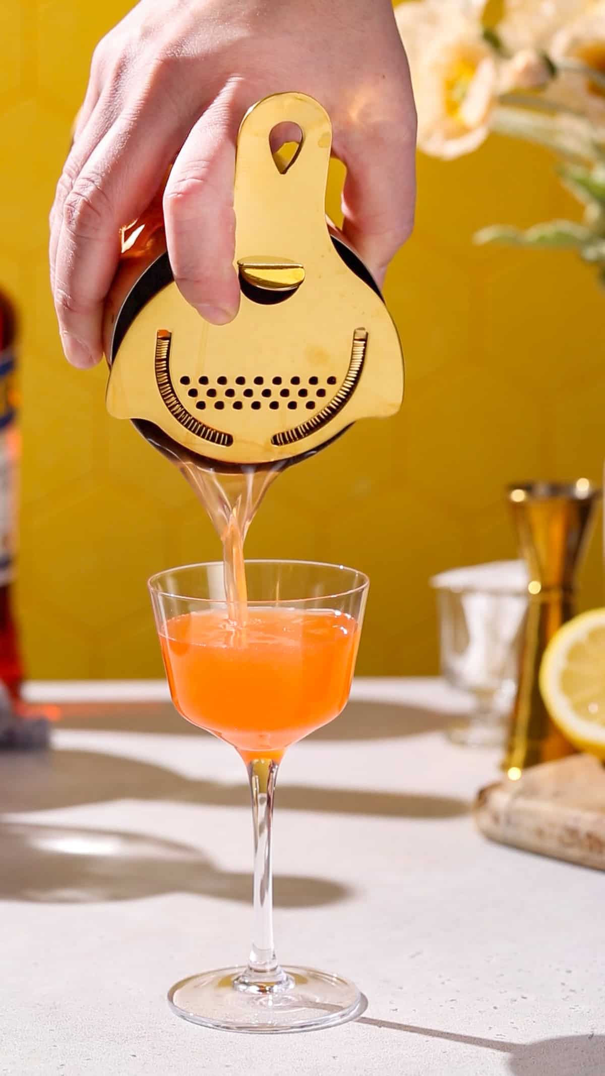 Hand using a gold cocktail shaker to strain an orange colored drink into a coupe glass.