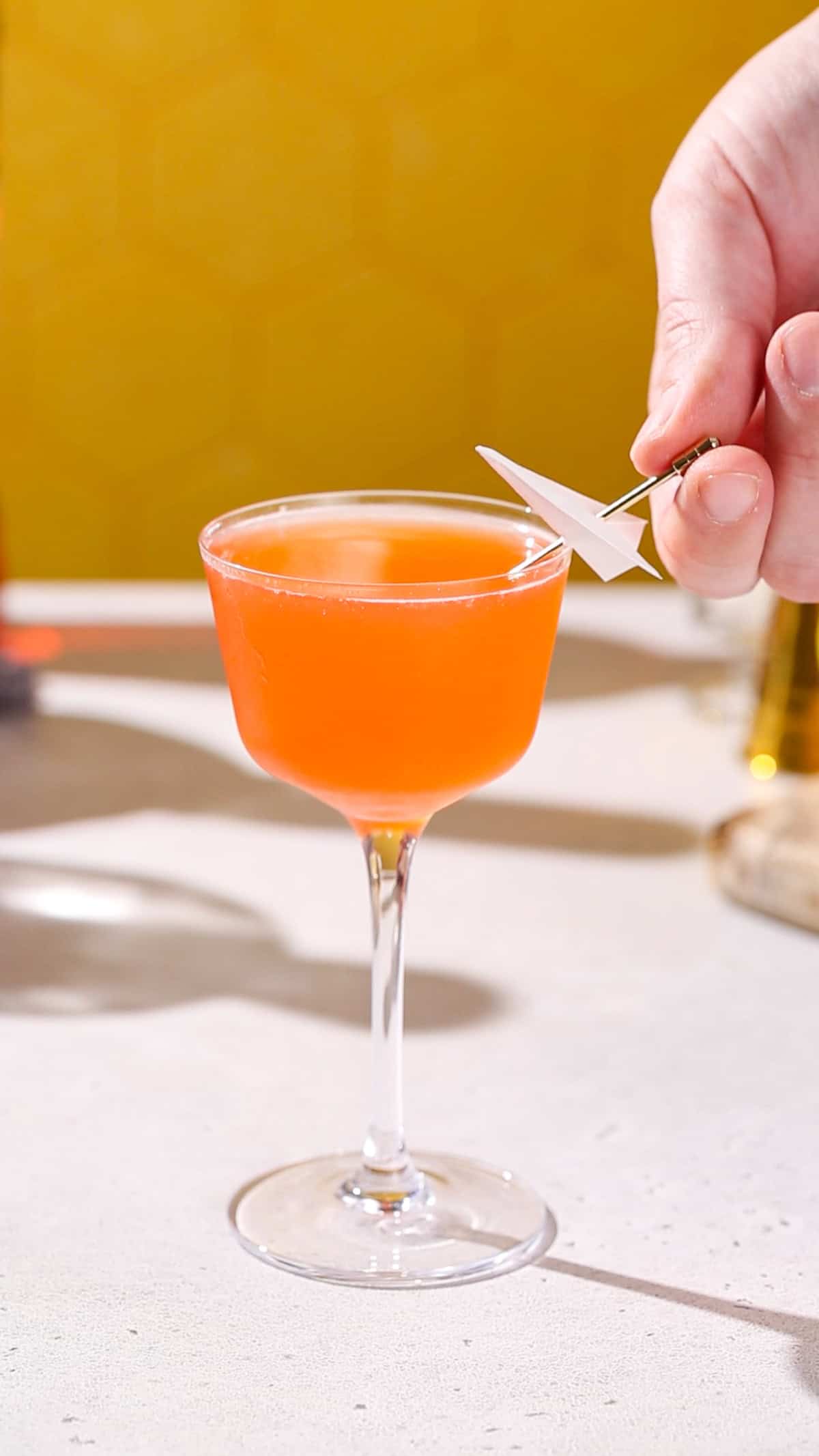 Hand adding a mini paper plane on a cocktail pick to an orange colored cocktail.