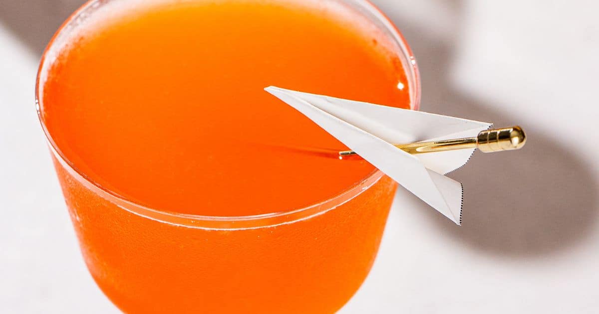 Close up of mini paper plane garnishing an orange colored cocktail.