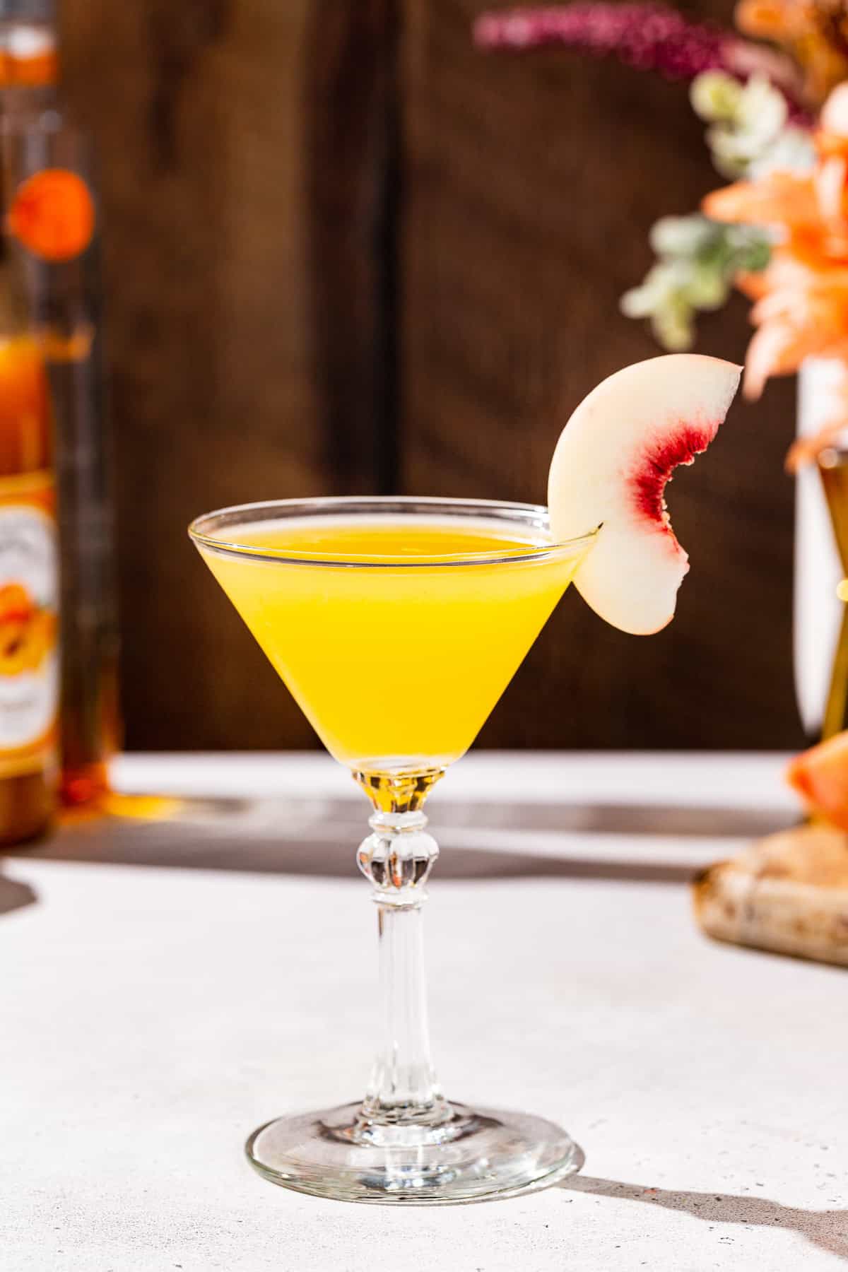 Side view of a Peach Martini cocktail on a countertop garnished with a peach slice. Ingredients and bar tools used to make the drink are in the background.