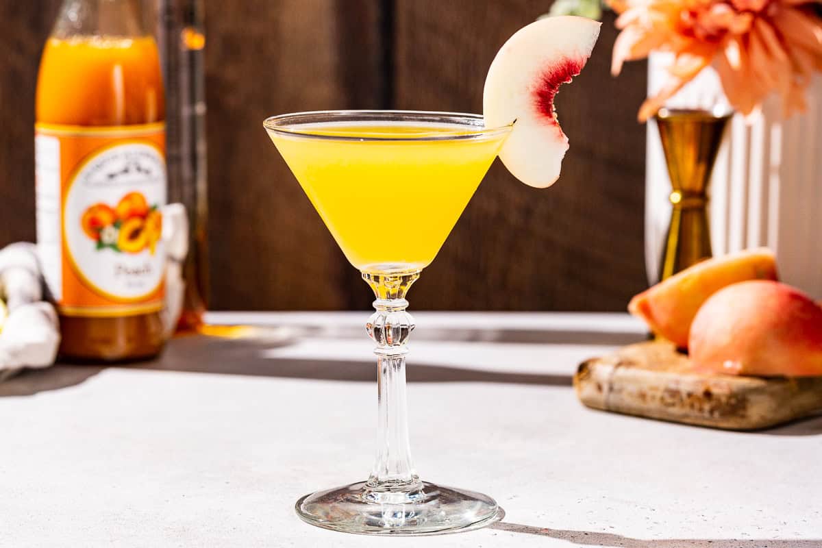 Side view of a Peach Martini cocktail on a countertop garnished with a peach slice. In the background are bottles of peach vodka and peach syrup on the left, as well as a fresh peach and a gold jigger on the right.