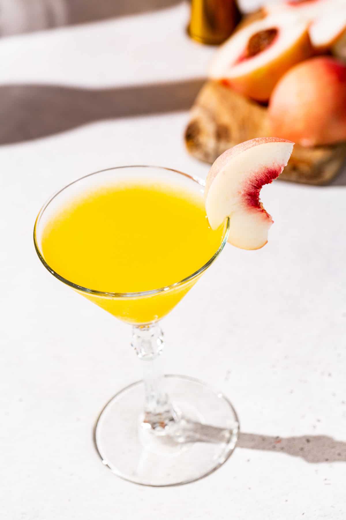 Overhead view of a Peach Martini cocktail. A fresh peach slice is a garnish and the rest of the peach is in the background on a cutting board.