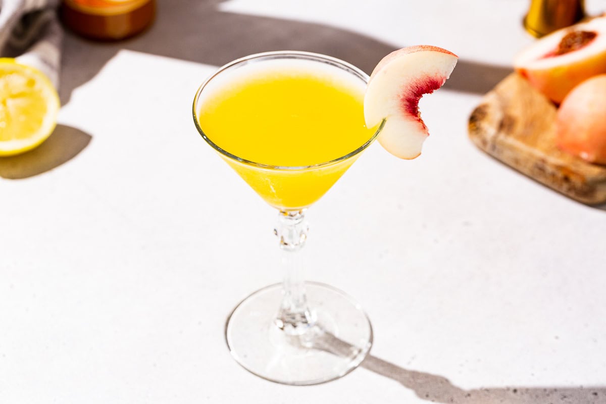 Peach Martini vodka cocktail on a countertop. In the background is a cut lemon and fresh peach.