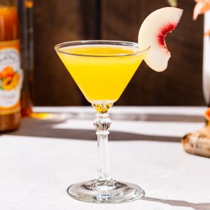 Peach Martini cocktail with some ingredients and bar tools used to make the drink in the background. The drink is a yellow color and is garnished with a slice of white peach.