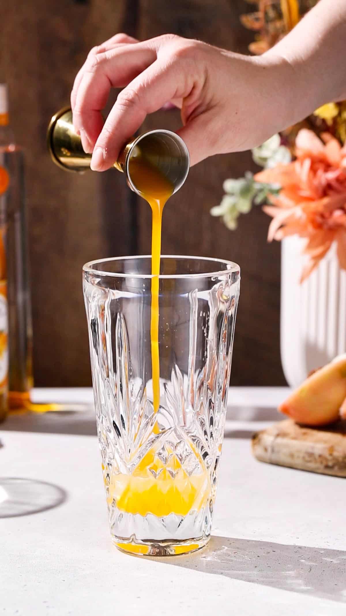 Hand using a jigger to add peach syrup to a cocktail shaker.