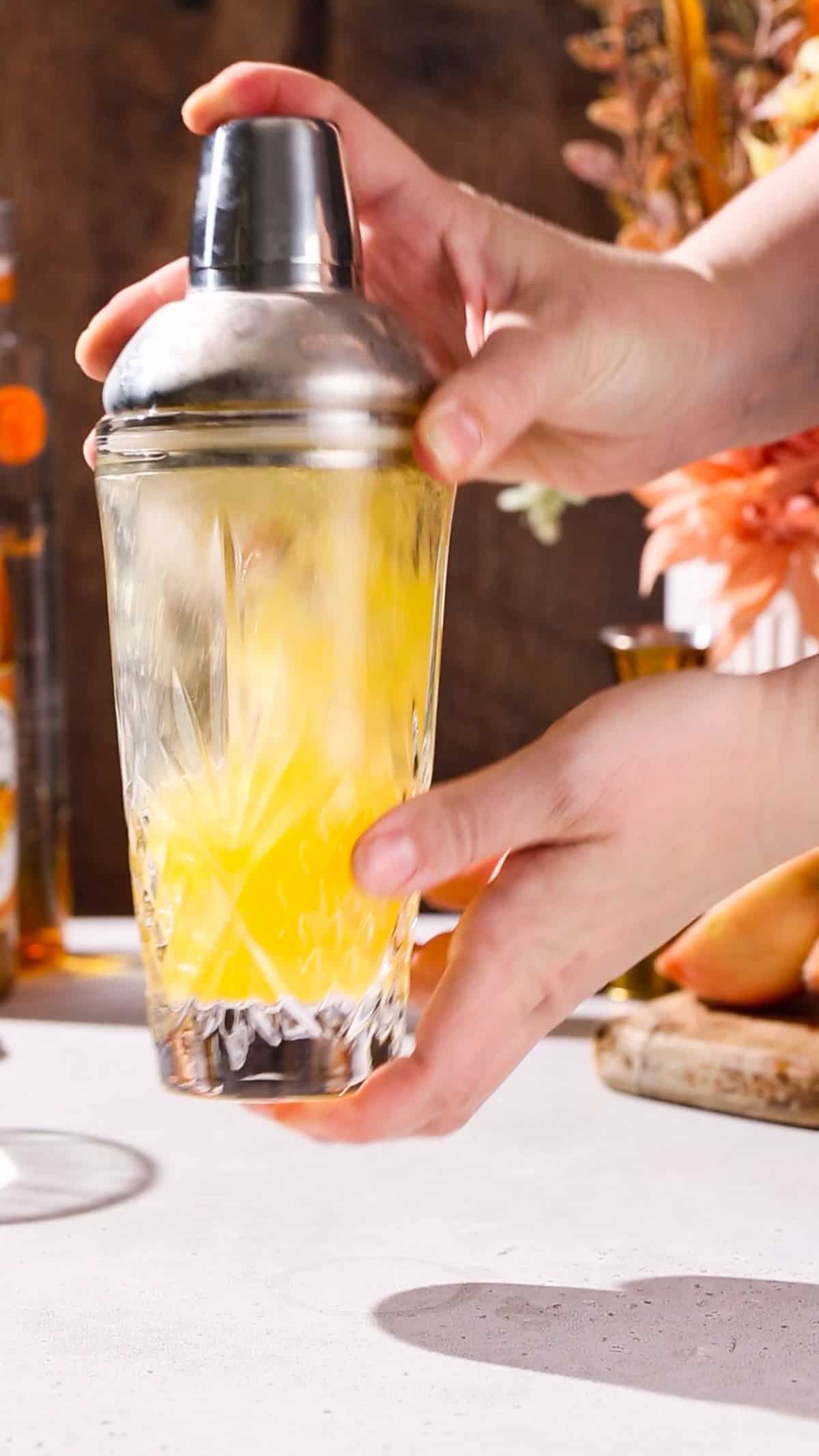 Hands shaking up a cocktail shaker filled with yellow liquid.