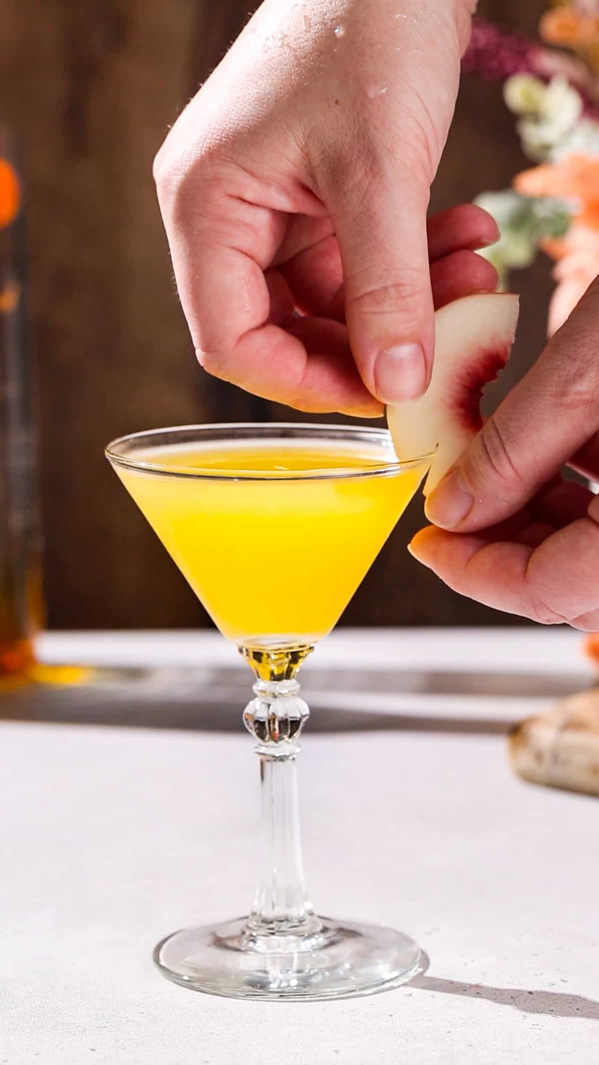 Hands adding a slice of peach to the rim of a martini glass filled with yellow liquid.