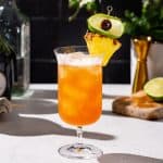 Side view of a Singapore Sling cocktail in a fancy serving glass. The drink is garnished with a pineapple, lime slice and cherry and there are some bottles and bar tools in the background of the image.