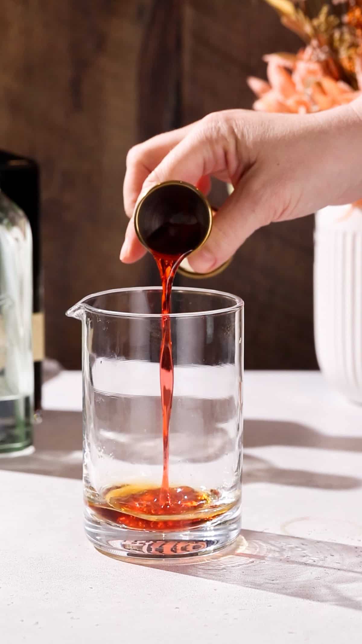 Hand pouring campari liqueur into a mixing glass.