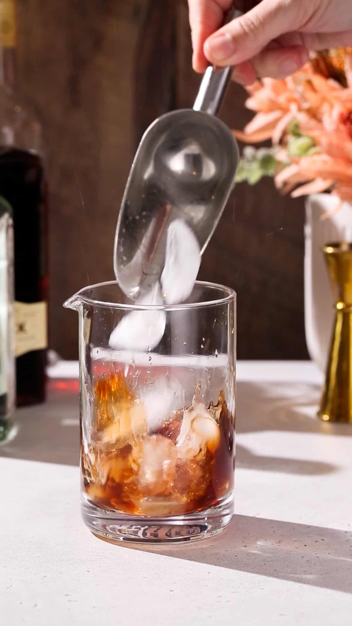 Hand adding ice to a cocktail mixing glass filled with brown liquid.