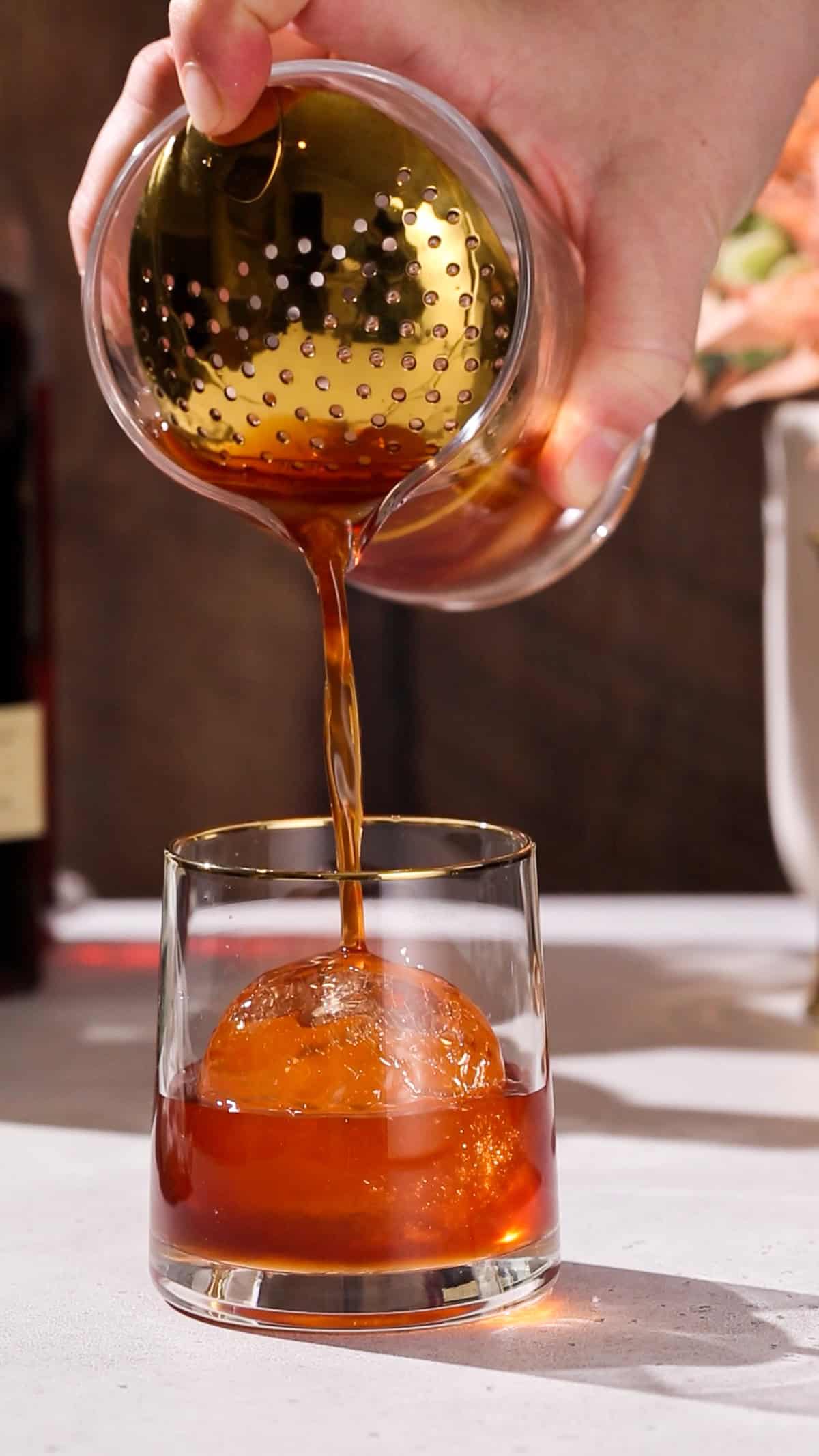 Hand straining a brown colored cocktail into a cocktail glass that has a large sphere of ice in it.