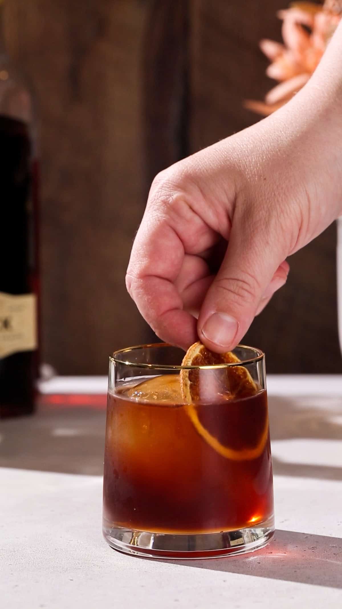 Hand adding a dried orange slice to a brown cocktail.