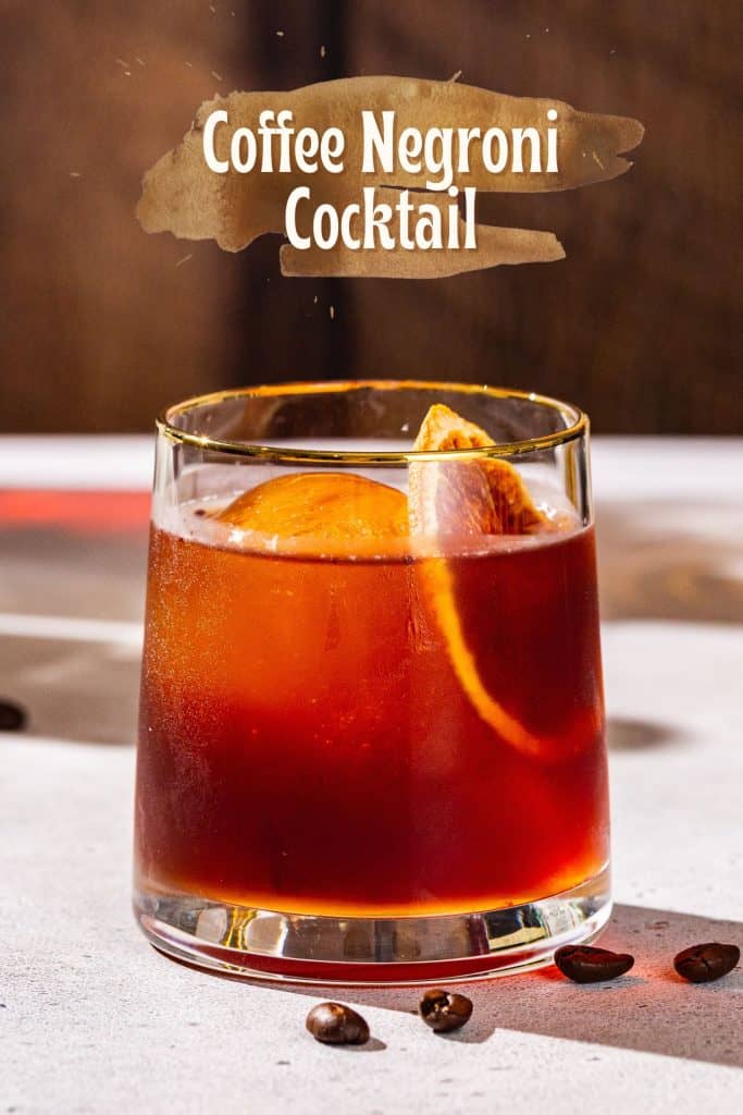 Coffee Negroni cocktail on a countertop with coffee beans scattered around it, garnished with a dried orange slice. Text above the drink says “Coffee Negroni Cocktail”.