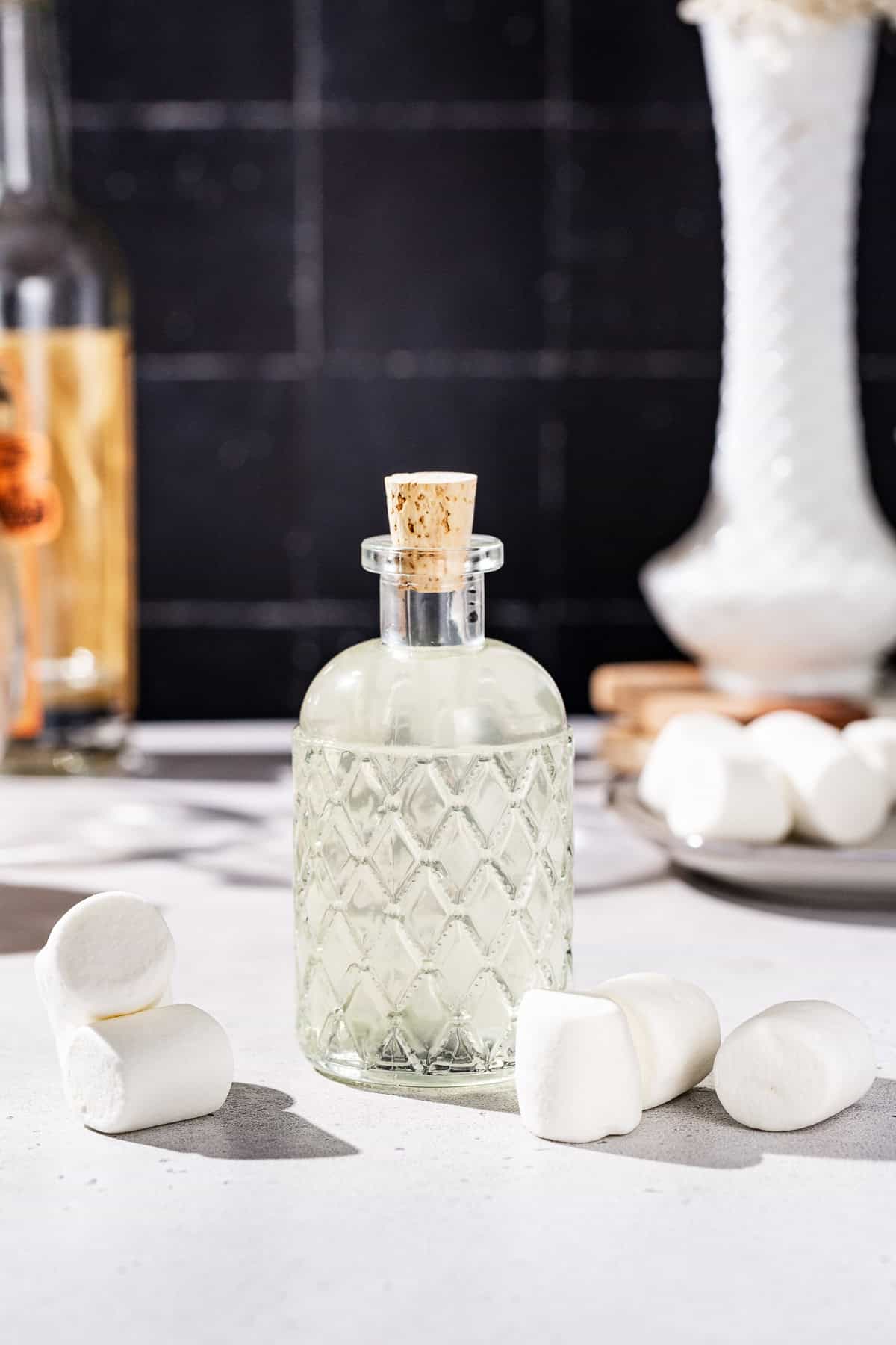 Side view of marshmallow vodka in a glass bottle, with marshmallows on the countertop around the bottle. There is a bottle of vodka and more marshmallows in the background.