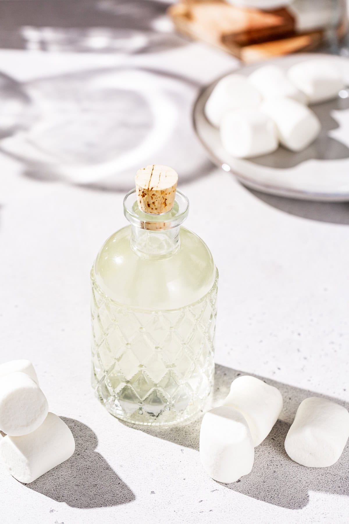 Glass bottle of marshmallow vodka on a countertop with marshmallows all around it. A dish of marshmallows is in the background.