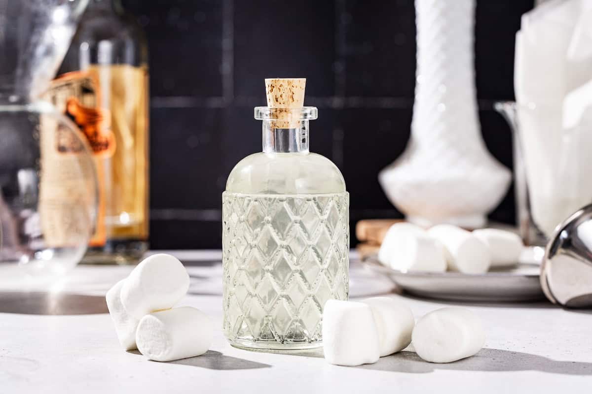 Glass bottle of marshmallow vodka on a countertop with marshmallows all around it. A dish of marshmallows is in the background.