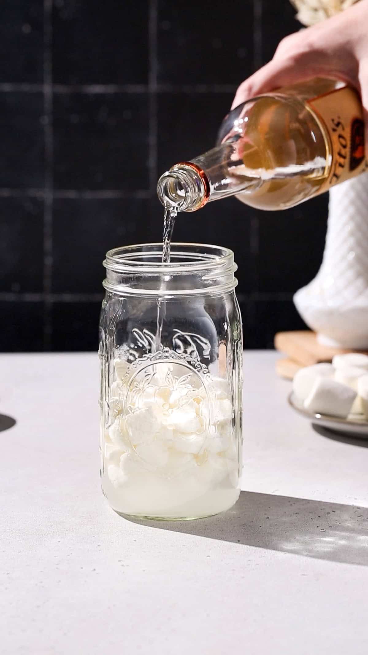 Hand pouring vodka into a jar filled with marshmallows.