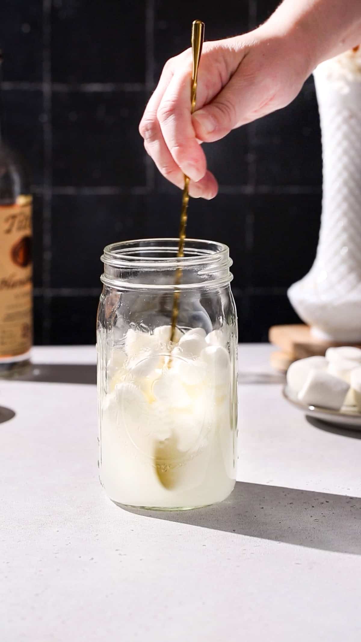 Hand using a long bar spoon to stir a jar of marshmallows and vodka.