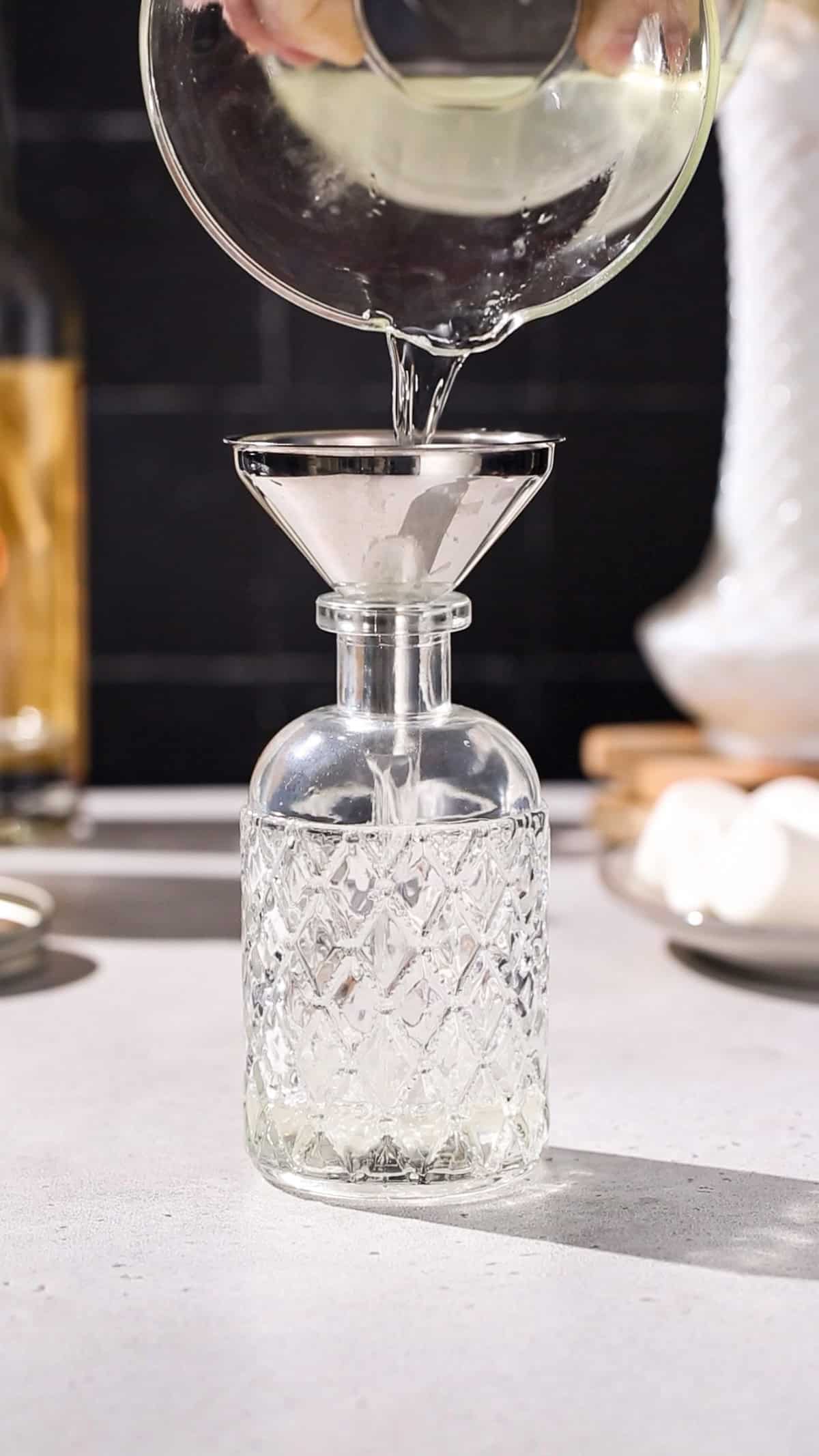 Hand pouring infused vodka from a carafe through a funnel into a glass bottle.