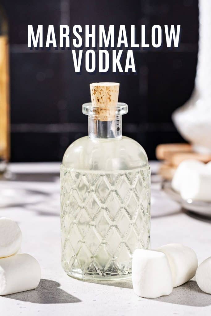 Bottle of homemade marshmallow vodka on a countertop surrounded by marshmallows. Text at the top says “Marshmallow Vodka”.