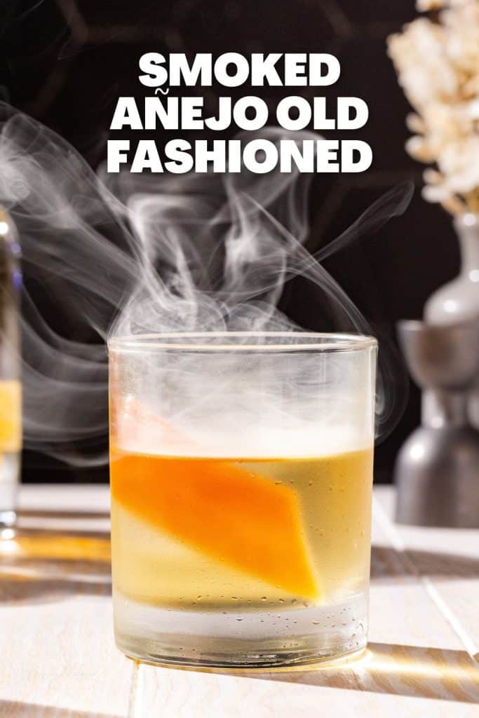 Side view of a Smoked Anejo old fashioned cocktail with a grapefruit peel garnish. Smoke is floating in and above the glass. Bold white text above the glass says “Smoked Añejo Old Fashioned”.