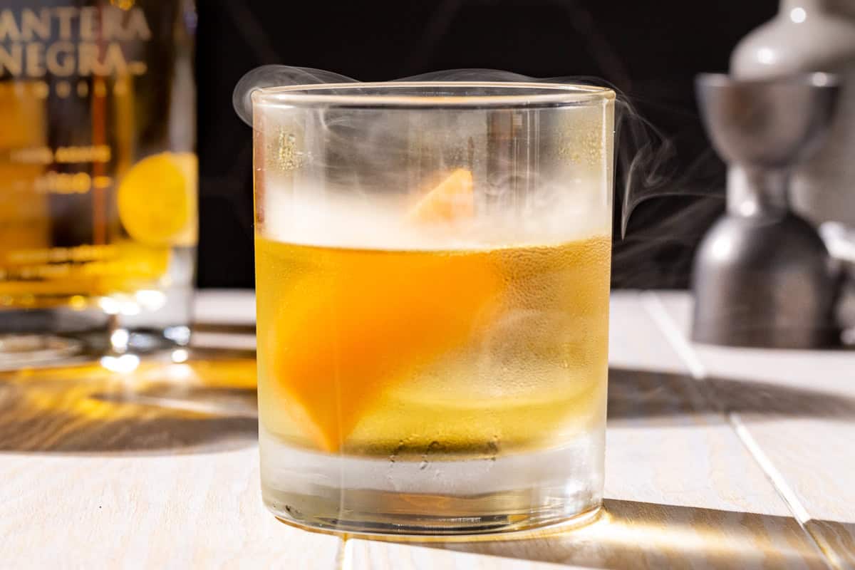 Side view of a Smoked Añejo Old Fashioned cocktail garnished with a grapefruit peel, with a bottle of anejo tequila in the background as well as a bell jigger.