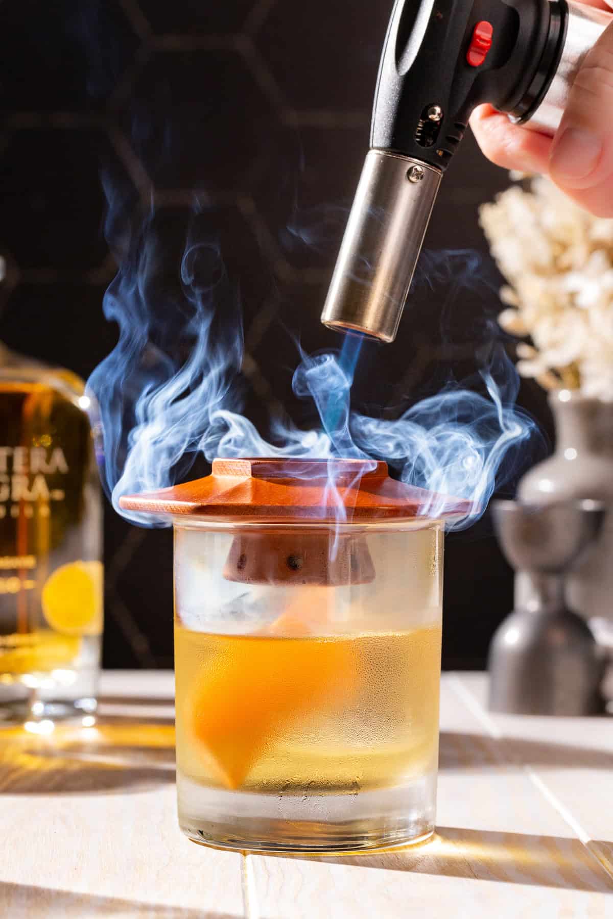 Hand using a kitchen torch to add smoke to a cocktail using a cocktail smoker.