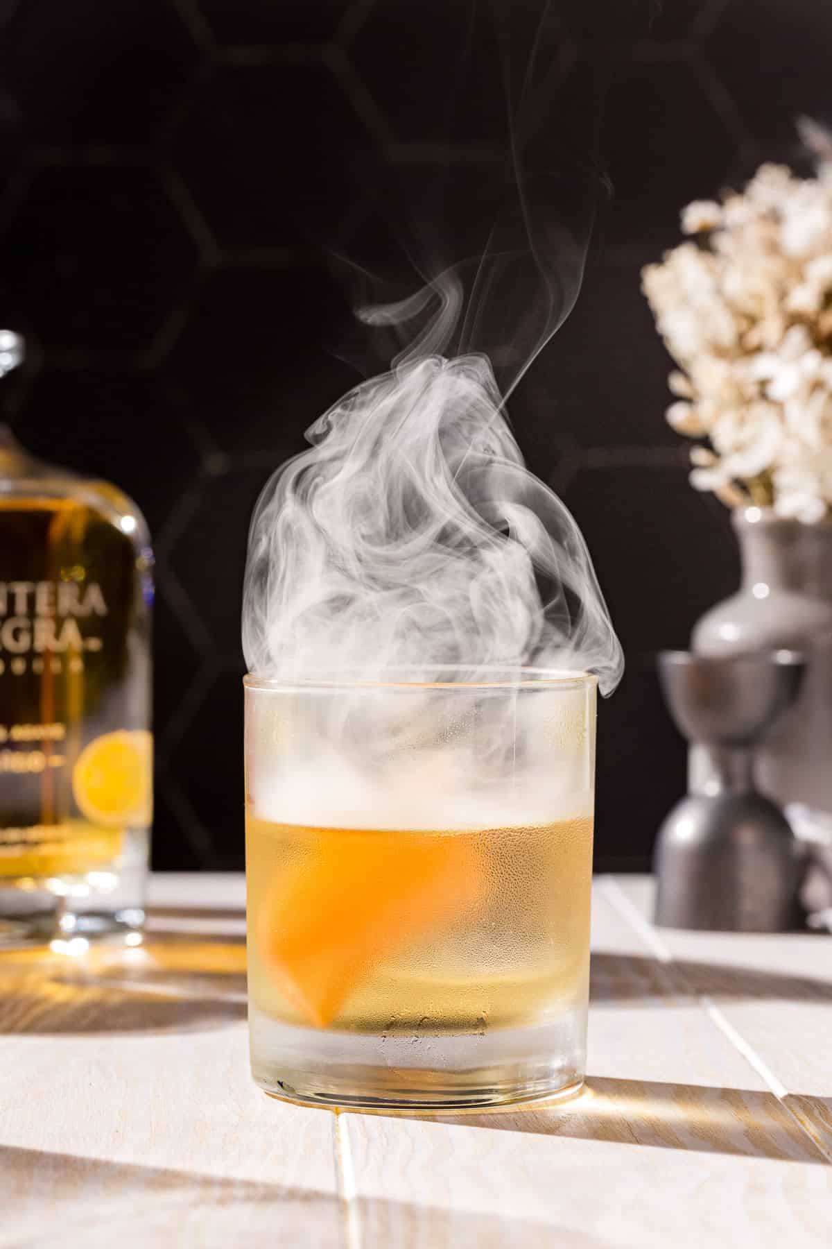 Smoked Añejo Old Fashioned cocktail with smoke coming out of the top and a grapefruit peel garnish.