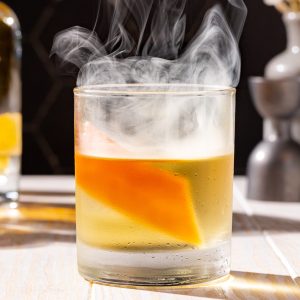 Smoked Añejo Old Fashioned cocktail with smoke coming out the top of the glass. The drink is garnished with a grapefruit peel.