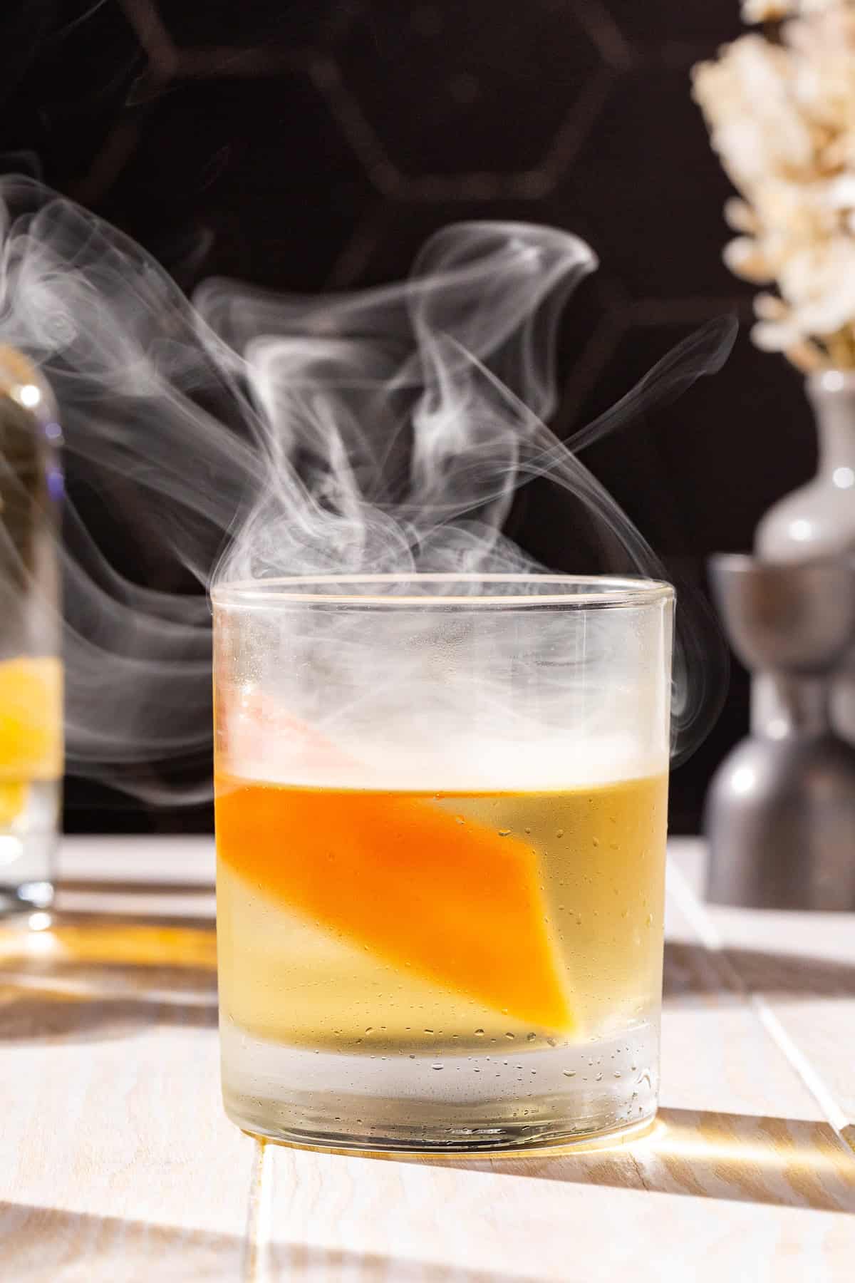 A Smoked Añejo Old Fashioned cocktail on a countertop with tequila and a jigger in the background. There is smoke coming up out of the glass.