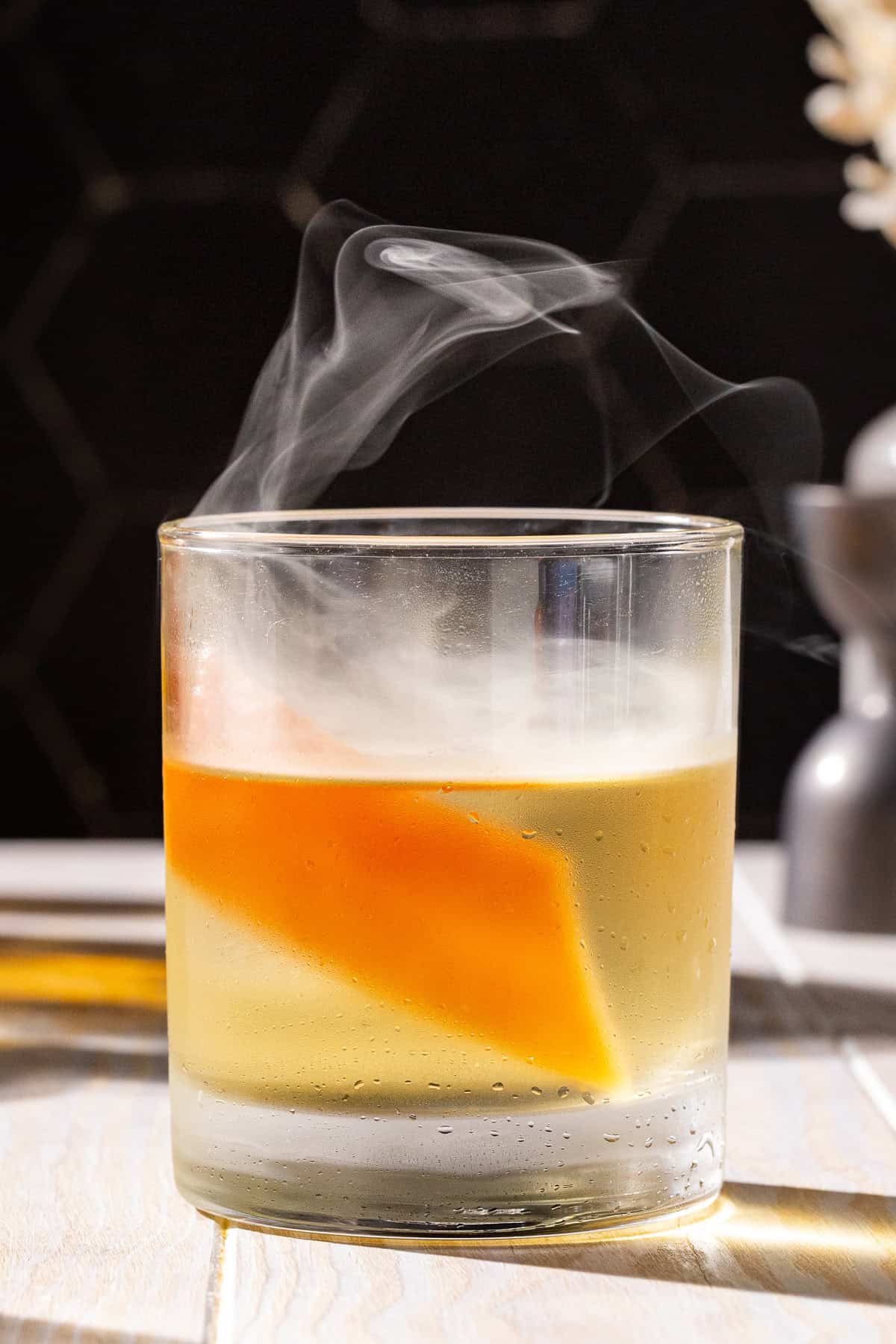 Side view of a Smoked Anejo old fashioned with a grapefruit peel garnish. Smoke is floating in and above the glass.