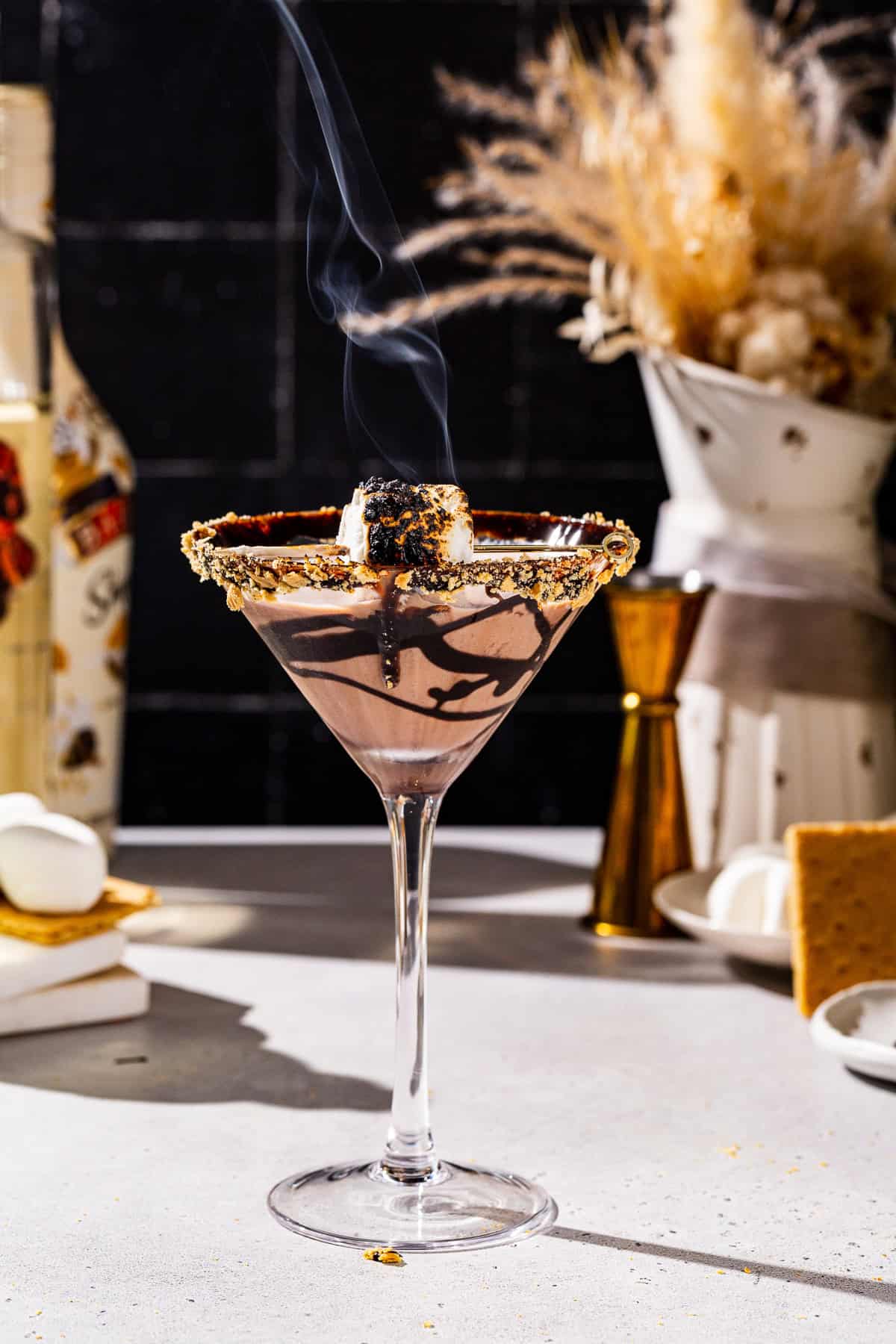 Side view of a S’mores martini cocktail in a tall stemmed martini glass. The drink is garnished with a toasted marshmallow, chocolate syrup and graham cracker crumbs on the rim of the glass. Smoke is coming off of the toasted marshmallow.