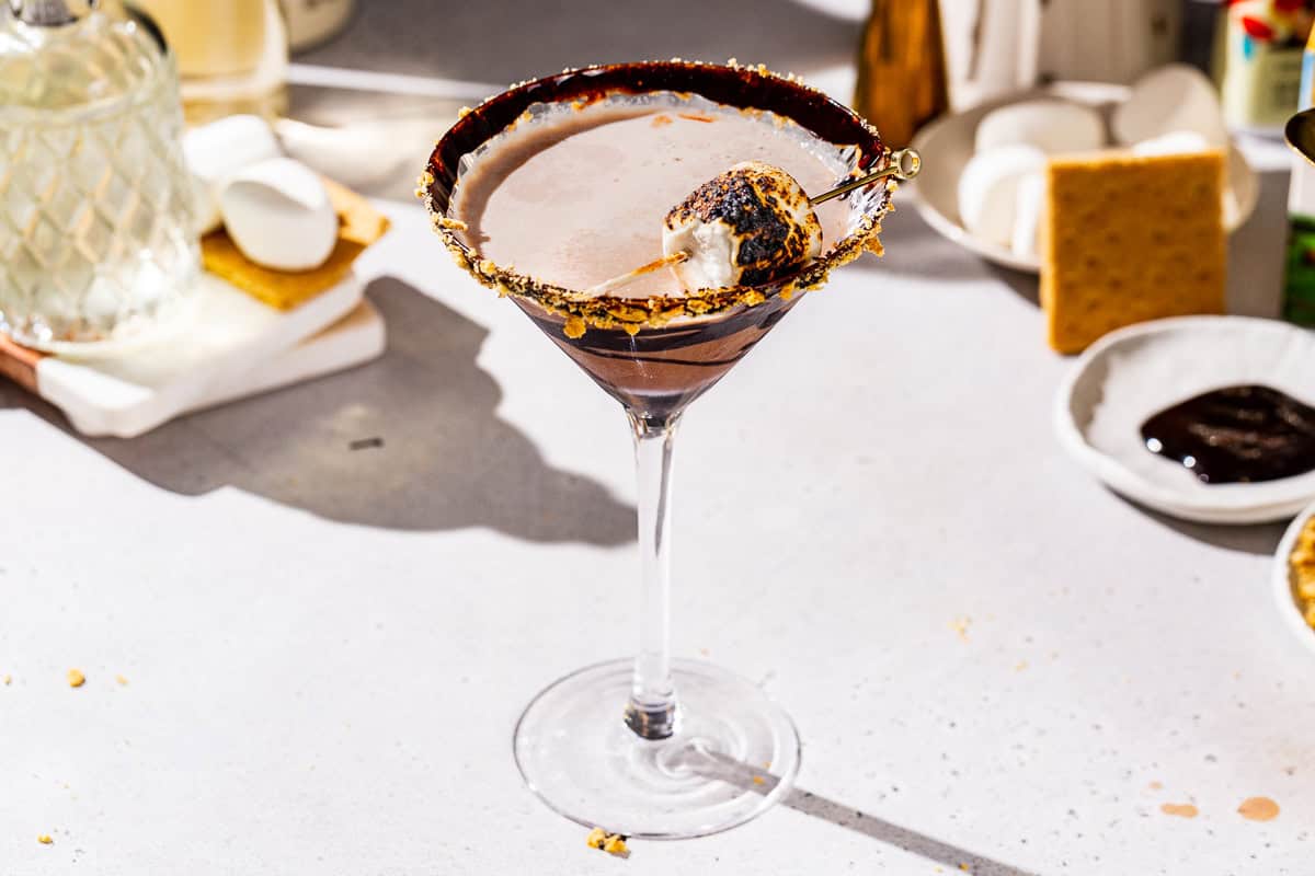 S’mores Martini on a countertop, garnished with graham cracker crumbs, chocolate syrup and a toasted marshmallow. In the background are some more marshmallows, graham crackers and other ingredients to make the drink.