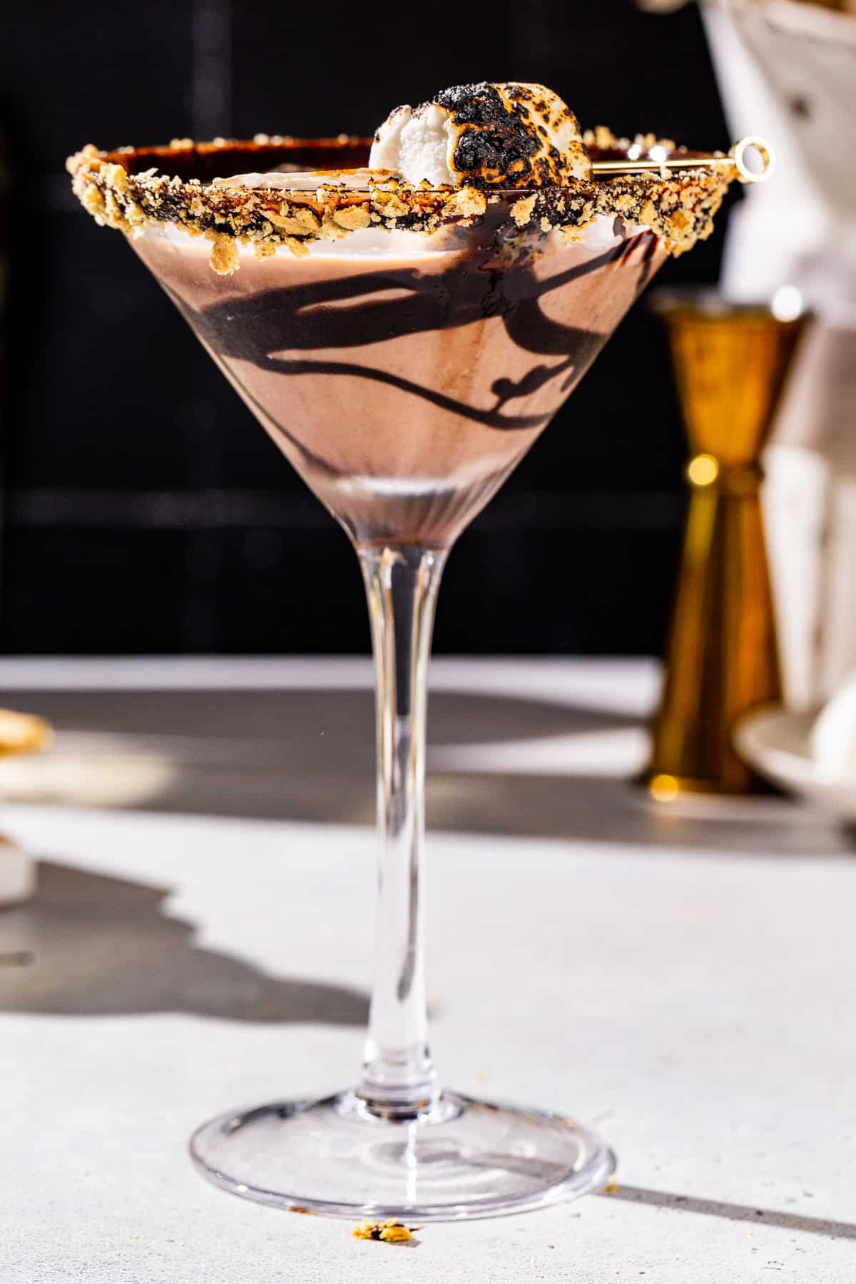 Side view of a smores martini. The drink is in a tall martini glass with a chocolate syrup drizzle, graham cracker crumbs on the rim and a toasted marshmallow on top.