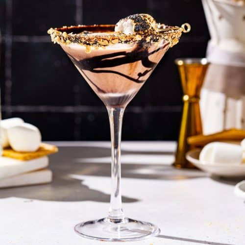 Side view of a smores martini. The drink is in a tall martini glass with a chocolate syrup drizzle, graham cracker crumbs on the rim and a toasted marshmallow on top. In the background is a jigger along with marshmallows and graham crackers.