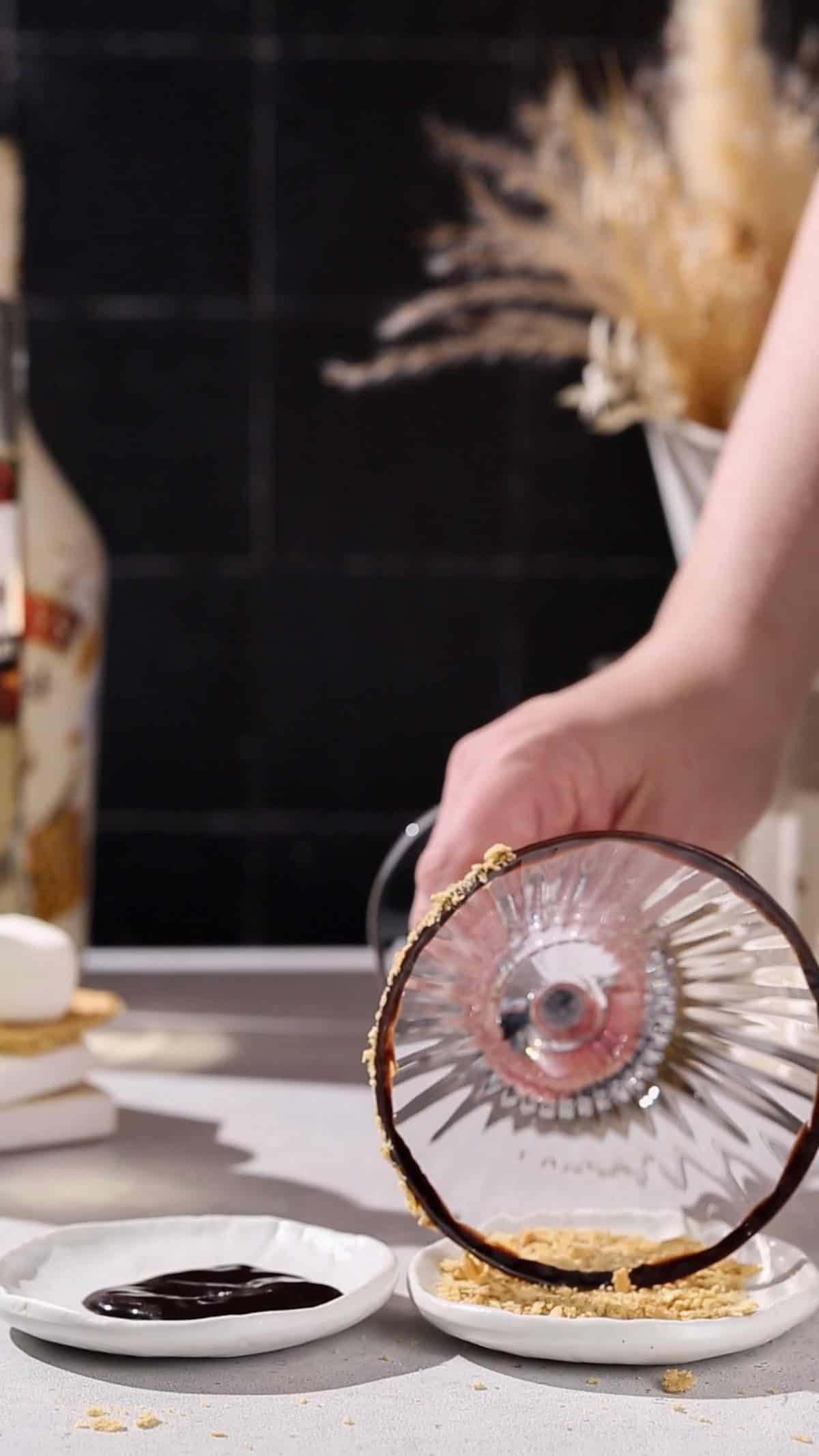 Hand dipping the rim of a martini glass into a dish of graham cracker crumbs.