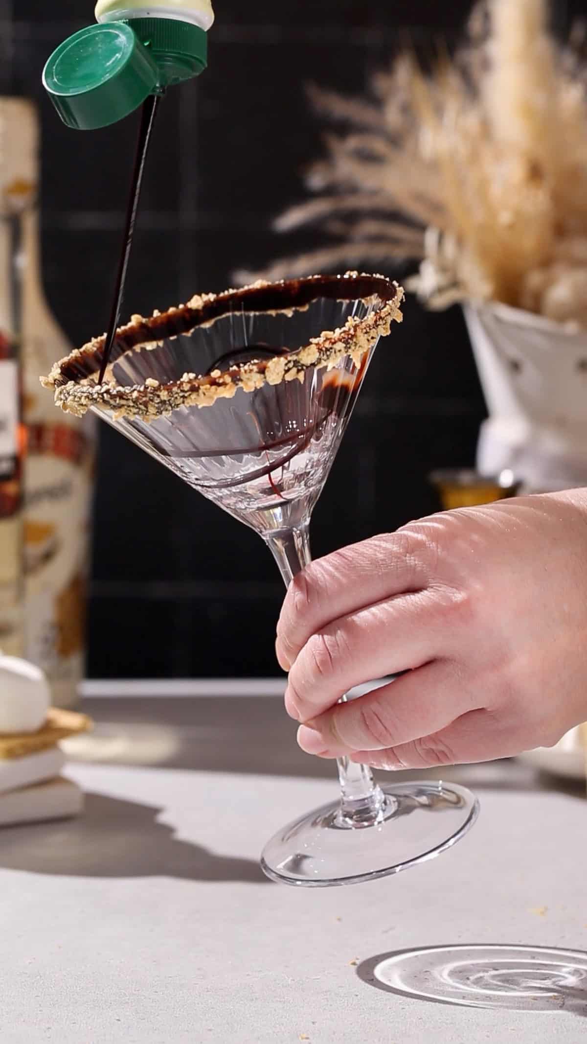 Hand adding a chocolate syrup drizzle to the inside of a cocktail glass.