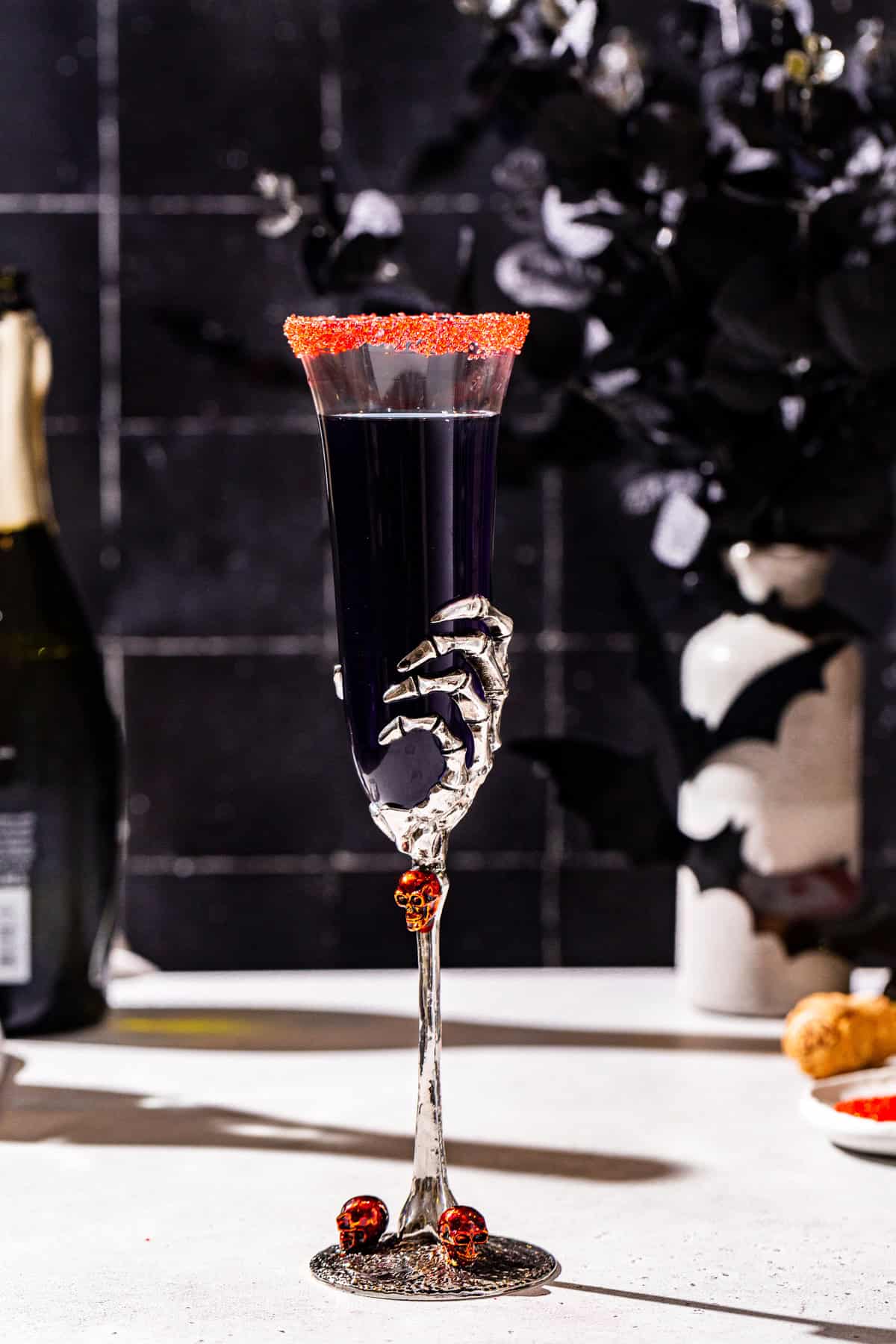 Side view of a Black Mimosa champagne cocktail with a red sugar rim. The drink is in a champagne flute with a skeleton hand for Halloween.