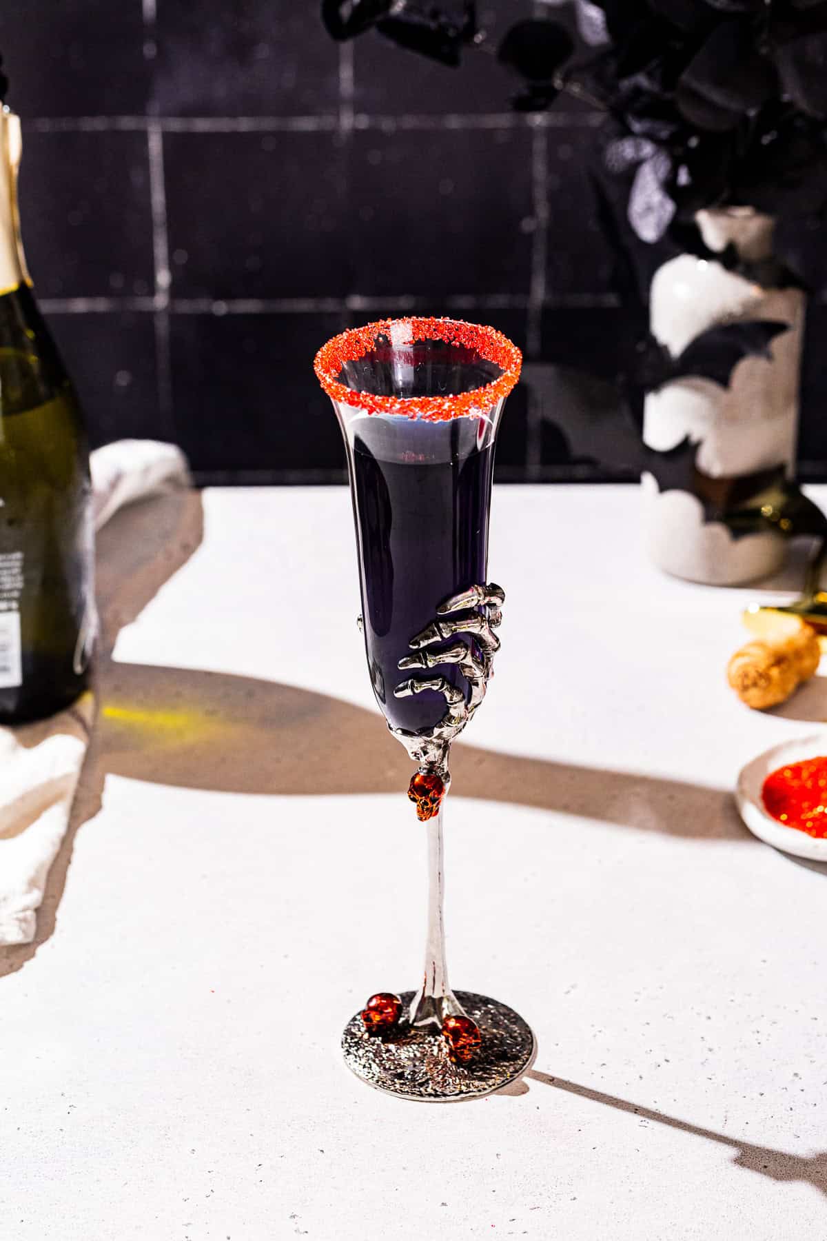 Black Mimosa champagne cocktail with a red sugar rim on a countertop with ingredients used to make the drink in the background.