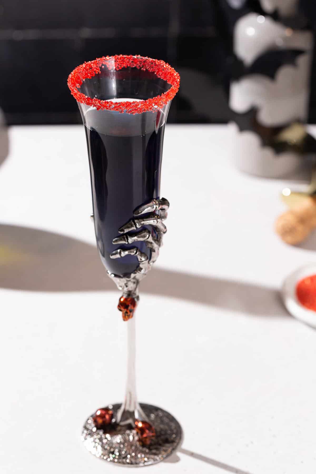 Black Mimosa cocktail with a red sugar glass rim. Ingredients used to make the drink as well as spooky Halloween decor is in the background.