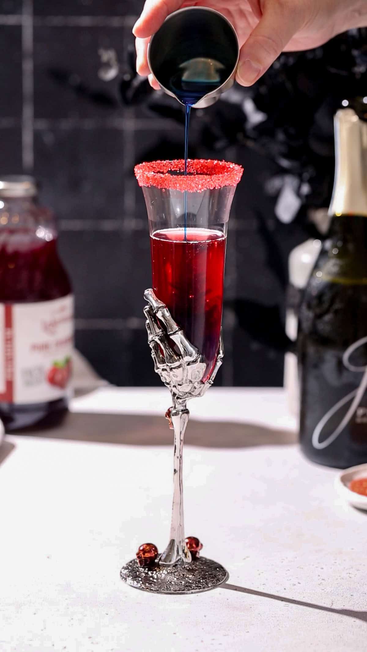 Hand pouring blue curaçao into a champagne flute filled with red liquid so it looks like the liquid is turning black.