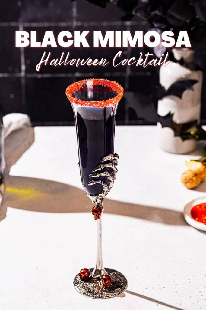 Black Mimosa cocktail in a champagne flute with a red sugar rim. The glass is a Halloween style glass with a silver skeleton hand. Text above the drink says “Black Mimosa Halloween Cocktail”.